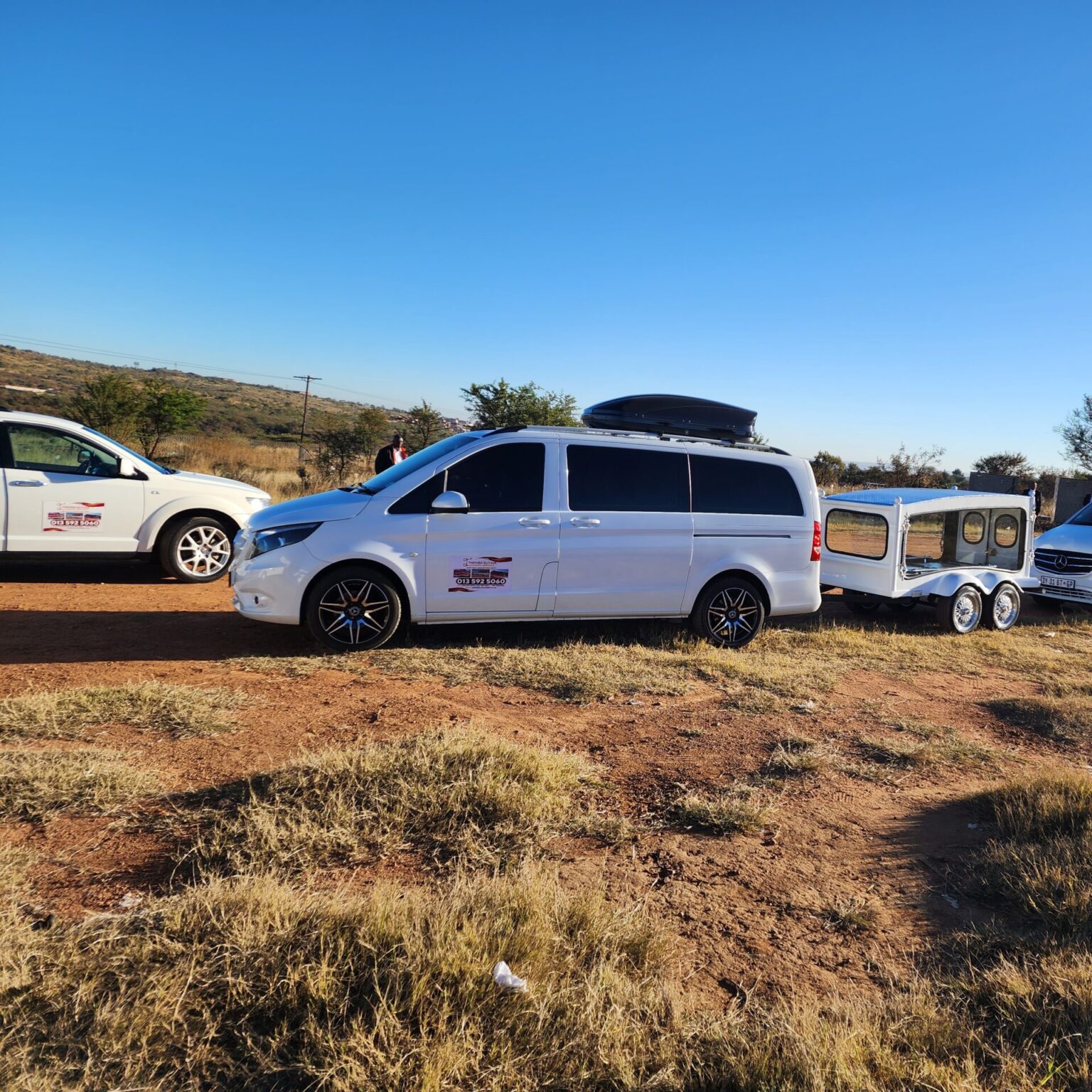 the image shows three vehicles belonging to Ithemba Elitjha Funeral services. The main vehicle in the picture is a white Mercedes Benz V-class branded with Ithemba Elitjha branding and behind it, there's a coffin carriage