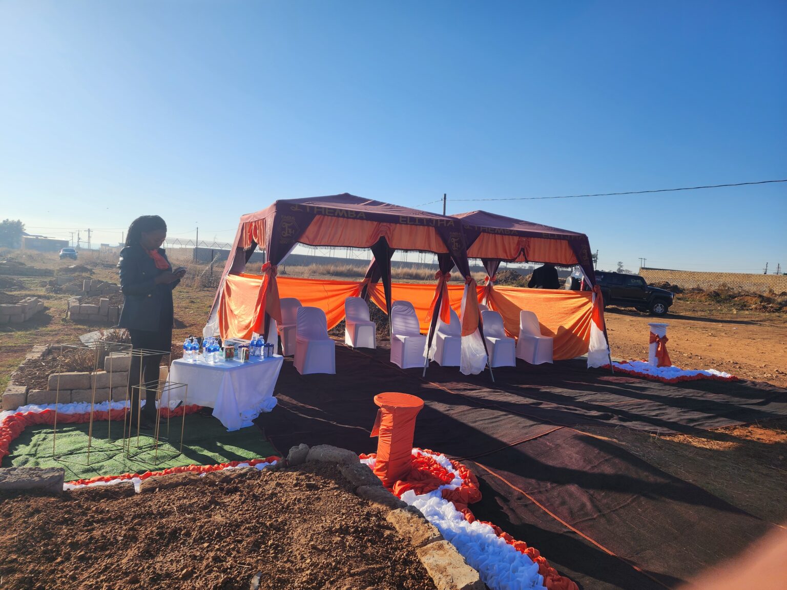 The image shows the graveside decor of Ithemba Elitjha Funeral Services, there are covered chairs under two gazebos that are branded with the Ithemba Elitjha name and logo, they are set up on a red carpet and there are orange and white pillars on the side.