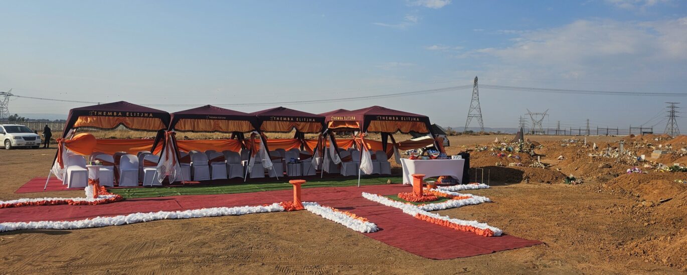 the image shows graveside Ithemba Elitjha deco. There are four maroon branded gazebos with a large number of white covered chairs underneath, all on the red carpet with orange and white grave side decorations.